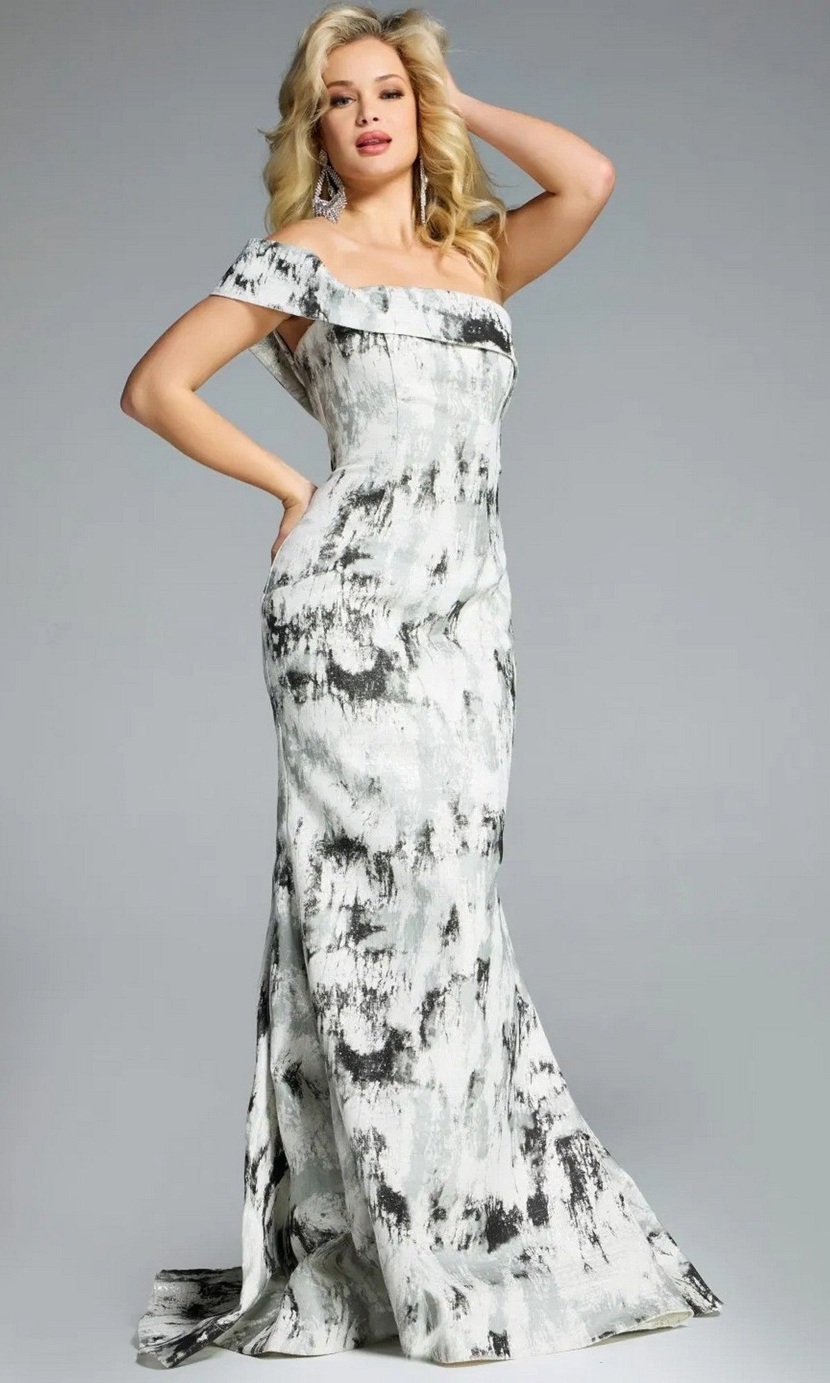 A woman poses in the Jovani 43977 long prom dress in grey, featuring an off-the-shoulder design and textured fabric, with one hand on her hip and the other touching her hair against a neutral background.
